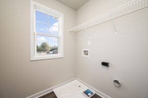 Empty laundry room with a window, wire shelf, and washer/dryer hookups on a light-colored wall.