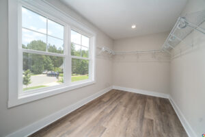 A spacious empty walk-in closet with light-colored walls, wooden flooring, and shelves on three sides. Sunlight streams through a large window overlooking a green outdoor area with trees and a street.
