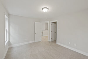 Empty room with beige carpet, white walls, and a ceiling light fixture. The room has a window on the left, a partially open door leading to a hallway, and electrical outlets on the right wall.
