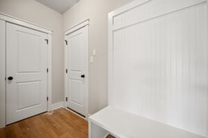 A small room with white walls, two white doors with black handles, and a built-in white bench with beadboard paneling beside one door. The floor is wooden.