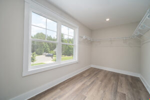 A walk-in closet with light wood flooring, beige walls, multiple white wire shelves, and a large window with a view of trees and a driveway.