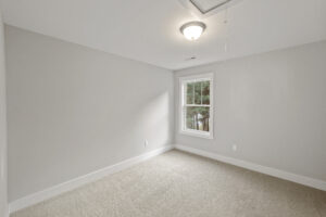 A small, empty room with light gray walls, beige carpet, a ceiling light, and a single window showing trees outside.