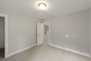 Empty room with beige carpet, light gray walls, a closed white door, a closet, a ceiling light fixture, and a visible staircase in the adjoining room.