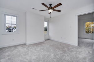 Empty room with white walls, gray carpet, a ceiling fan, two windows, and a doorway leading to another room. House and trees are visible outside the windows.