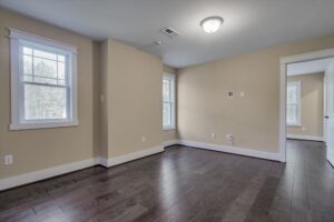 An empty room with beige walls, dark hardwood floors, two windows on one side, and a white door on the opposite wall.