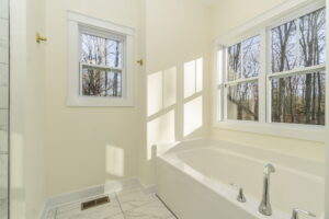 A bright bathroom with a soaking tub under large windows showing a view of leafless trees, white walls, and a tiled floor.