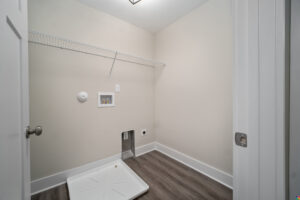 A beige laundry room with a wire shelf, hookups for washer and dryer, an empty washer tray on the floor, and hardwood flooring.