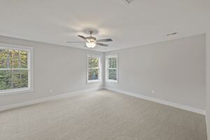 A spacious, empty room with beige carpet, white walls, a ceiling fan with lights, and two double windows letting in natural light.