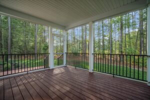 A screened-in porch with wooden flooring and black metal railings, overlooking a forested backyard area.