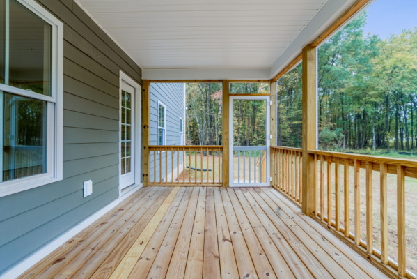 Wooden porch with railing, leading to a grassy yard with trees in the background. The porch is connected to a house with gray siding and a glass door.