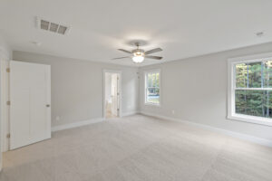 Spacious empty bedroom with light gray walls, a white ceiling fan, and two large windows. There's a door leading to an adjoining bathroom. The room is carpeted in a light beige color.