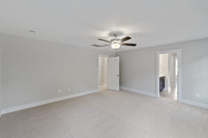 A spacious, empty room with light-colored carpet, gray walls, a ceiling fan with light, a white door slightly ajar, and an open doorway leading to another room with tiled flooring.