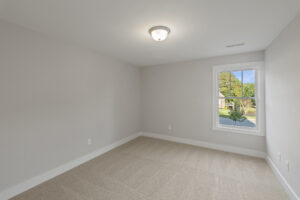 An empty, carpeted room with beige walls, a ceiling light fixture, and a window showing a view of greenery outside.