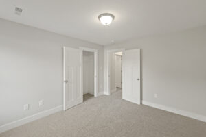 A clean, empty bedroom with carpeted floor, white walls, and a ceiling light. There are two open doors revealing closets and an adjacent room.