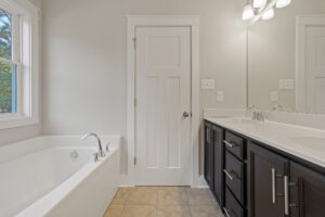 A bathroom with a bathtub, a sink with a large mirror, dark cabinets, a door, and white walls and trim.