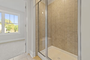 A bathroom with a glass-enclosed shower featuring beige tiles, adjacent to a room with a large window overlooking the outdoors.