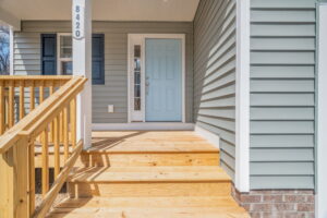 A light blue front door of a house with gray siding, wooden porch, and number 8420 visible next to the door under a clear sky.