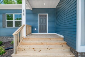 Exterior view of a house entrance with a gray door and blue siding, featuring a new wooden porch and steps.