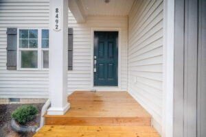 A front porch with light wooden steps, white siding, a dark green door, and gray shutters. The house number 8492 is displayed on a white column.