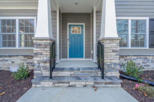 Front entrance of a modern house featuring a blue door, stone columns, and a small porch with railings.
