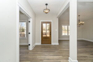 Modern empty room with wooden floor, white walls, and a wooden door at the center, leading outside; visible in daylight.