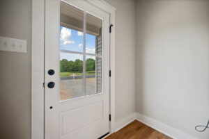 A white door with a window and black handles opens to a view of trees and a gravel driveway.