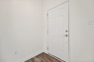 A white door with a round doorknob and deadbolt lock stands closed in a room with white walls and dark wood flooring. An electrical outlet and a light switch are visible on the walls.
