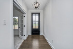 A narrow hallway with hardwood floors features a black front door with glass panes. To the left, there is a room with French doors and a carpeted floor. A modern hanging light fixture is overhead.