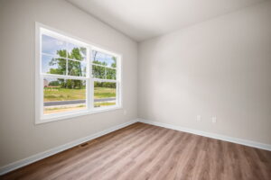 Empty room with beige walls, wooden floor, and a large window showing a view of green trees and a grassy area.