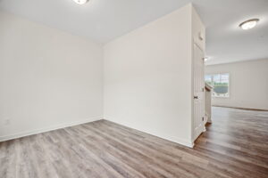 A spacious empty room with white walls, wooden flooring, a closed door, and a window letting in natural light.