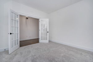A room with light gray carpeting, white walls, and double glass French doors opening into another room with wooden flooring and a hanging light fixture.