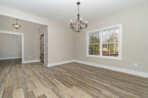 Empty living room with wooden floors, large windows, and modern chandeliers, facing a wooded outdoor area.