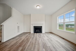 A living room with light-colored walls, wooden flooring, a fireplace, and a large window offering a view of houses and greenery outside. Stairs are visible on the left side.