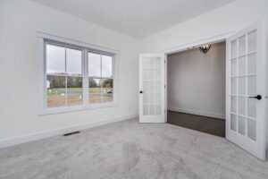 A bright, empty room with white walls and carpeted floor, featuring large windows and French doors leading to an adjacent space.
