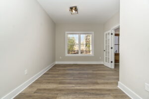 Empty room with wooden flooring, featuring a large window and a closed white door on the right. the walls are painted white.