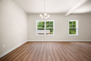 Empty room with beige walls and hardwood flooring featuring a chandelier and three windows, overlooking greenery outside.