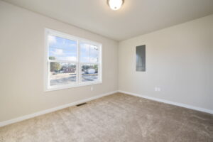 Empty carpeted room with a large window, beige walls, and an electrical panel on the right wall.