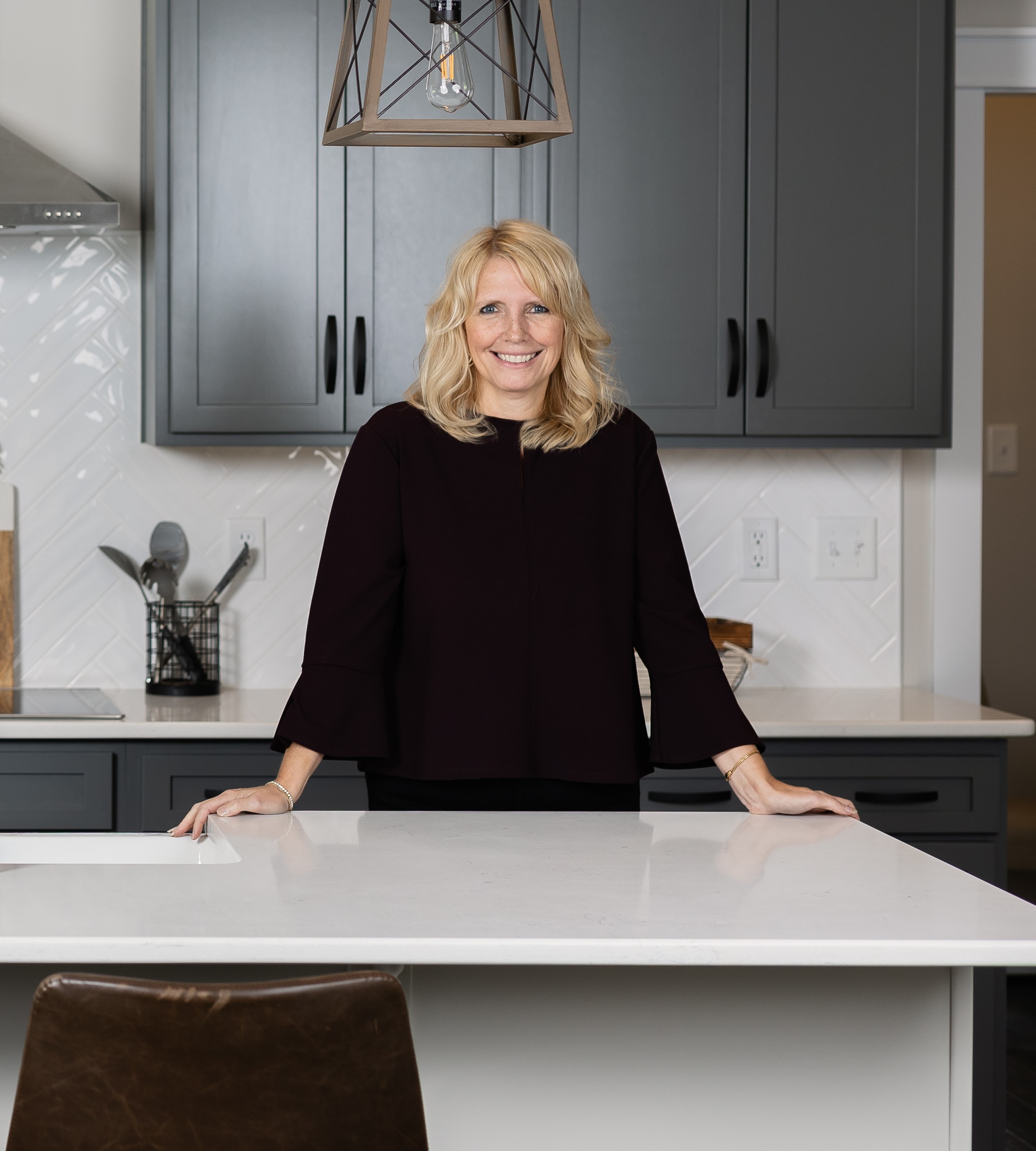 A woman with blonde hair stands behind a white kitchen island, resting her hands on it, with gray cabinets and a backsplash in the background.