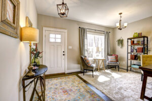A cozy, well-lit living room with two chairs, a round table, a bookshelf, and a door. The room has a rug, lamps, and wall decor, with sunlight streaming in through the window.