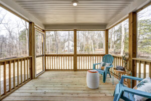A screened-in porch with wooden flooring featuring two blue chairs, a small table, and a view of a house and trees in the background.
