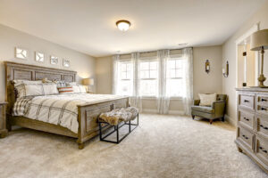 A cozy bedroom with a wooden bed, plaid bedding, fur bench, armchair, dresser, and large windows with sheer curtains. Walls are light beige with four framed decorations above the bed.