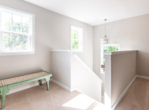 Bright, empty room with cream walls, carpeted floor, two windows, and a green bench. staircase with white railing visible on the right.