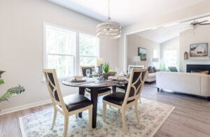 A bright, modern dining room with a set table for four, large windows, and an open view into a living room with a gray couch.