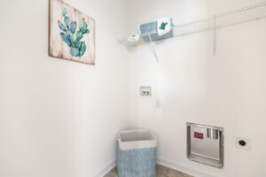 A small laundry room with white walls featuring a cactus painting, a shelf with detergent, a laundry basket, and a built-in wall chute.