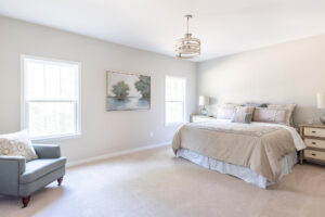 A bright and neatly arranged bedroom featuring a double bed with beige linens, a gray armchair, two windows, and a landscape painting on the wall.