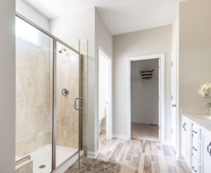 Modern bathroom interior with glass shower enclosure, beige tiles, white vanity, and open doors leading to adjacent rooms.