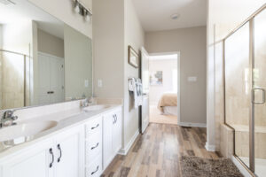 A modern bathroom with dual sinks, a large mirror, and a glass shower door leading to a bedroom visible in the background.
