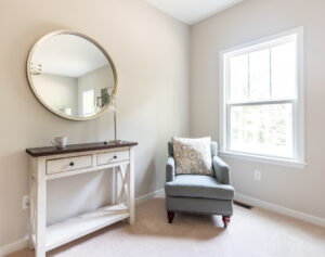 A bright, minimalist room featuring a white desk with a round mirror, a blue armchair with a decorative pillow, and a window showing greenery outside.