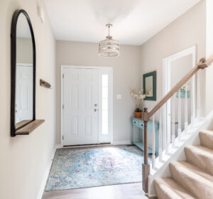 A bright, welcoming entryway with a white door, a staircase on the right, a decorative rug, and a mirror on the left wall.
