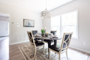 A bright dining room featuring a modern table set for four with plates and mugs, a decorative rug, large windows, and a pendant light.